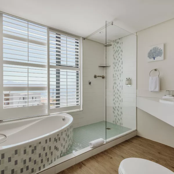 spacious hotel bathroom with spa bath, open shower, and large shuttered windows offering sea views.
