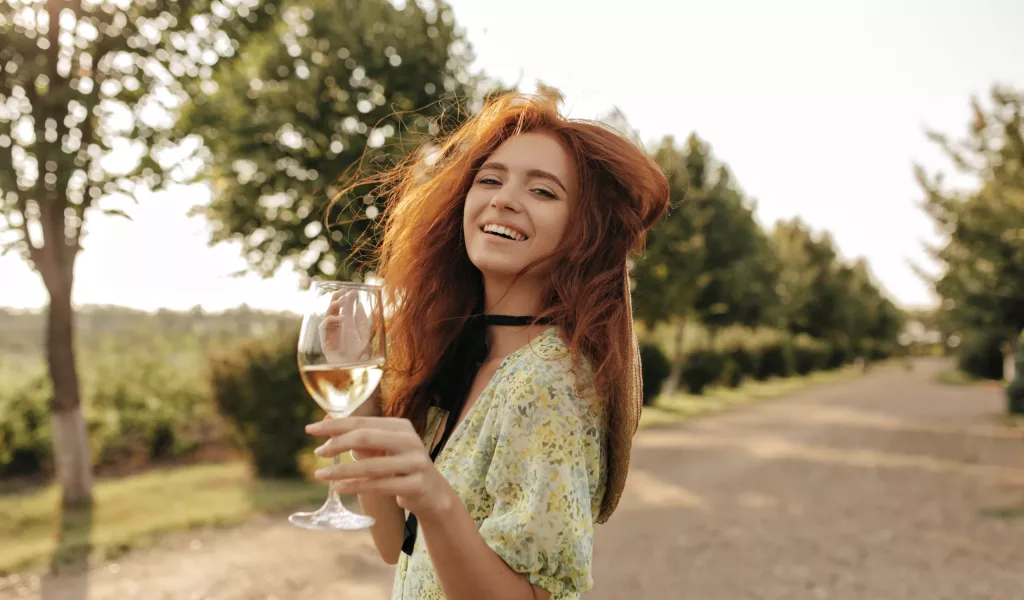 Happy girl with long ginger hair in summer yellow outfit smiling, looking into camera and holding glass with champagne outdoors